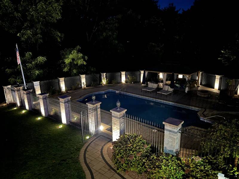 Elevated view of a pool and patio surrounded by illuminated stone fence pillars