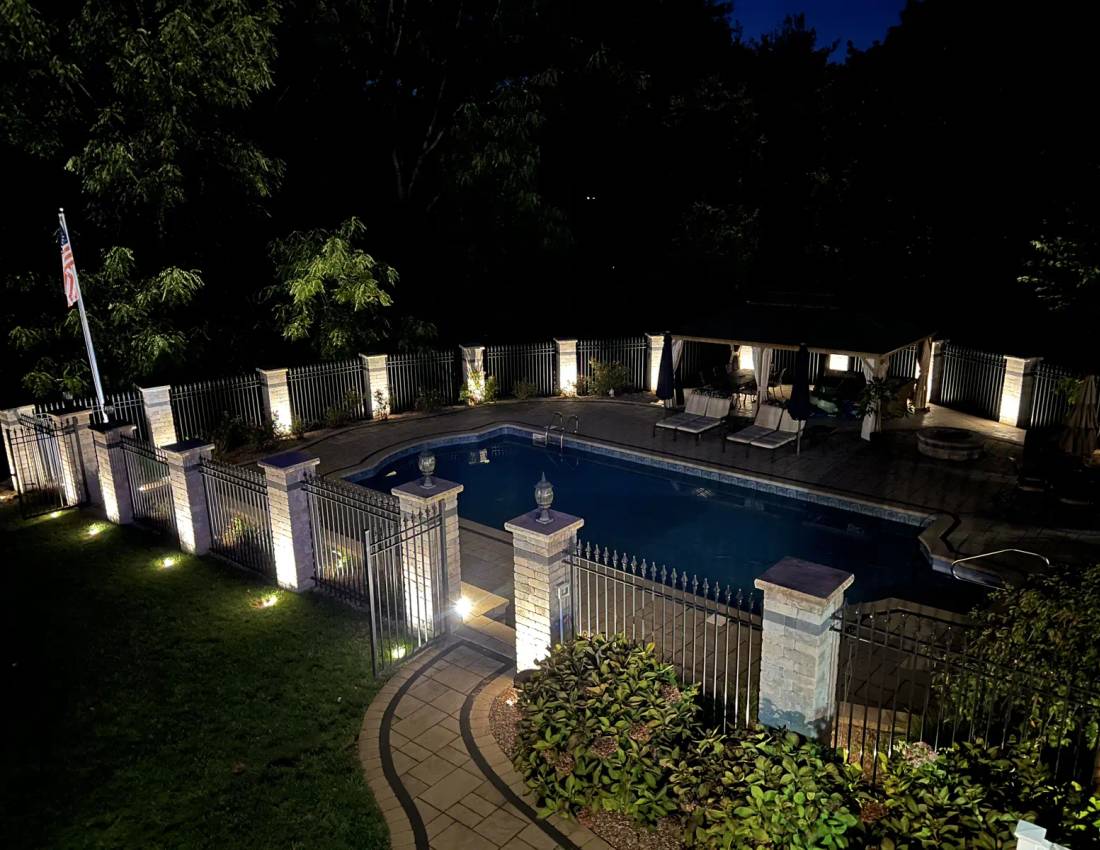 Elevated view of a pool and patio surrounded by illuminated stone fence pillars