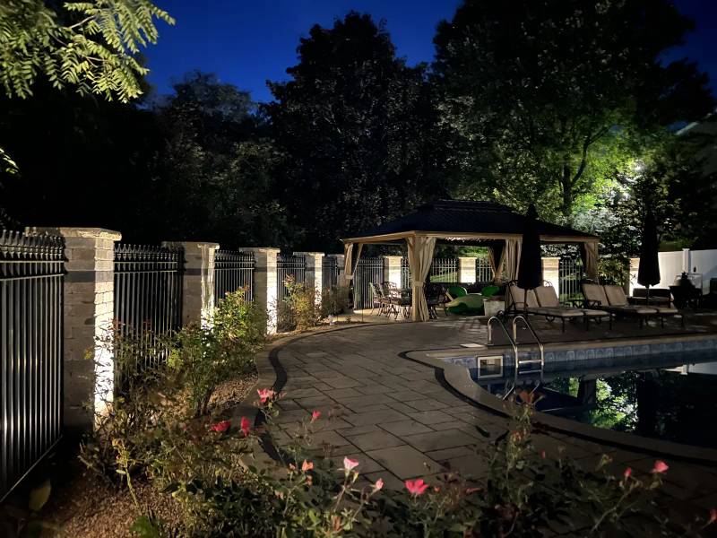 Lit stone pillars and garden beds along a poolside path leading to a pavilion